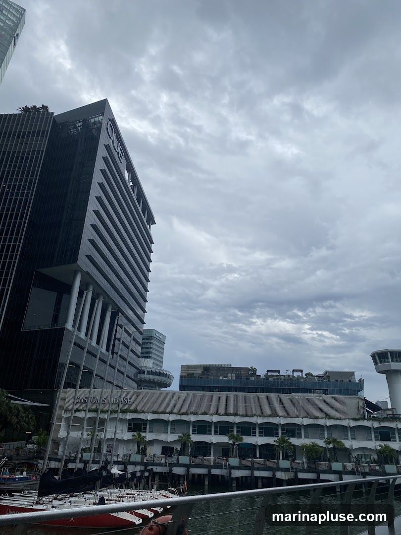 A view of modern glass-fronted buildings and a white, multi-story structure with a waterfront promenade. The sky is overcast with dramatic clouds. Boats are docked in the foreground. The word "QUE" is visible on the top of one building, and "CUSTOMS HOUSE" is visible on the white building.
