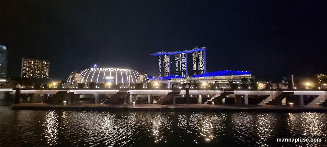 A nighttime view of The Fullerton Pavilion, a domed structure with illuminated ribs, situated by a body of water. In the background, the iconic Marina Bay Sands hotel complex is visible, lit up with blue lights. The scene is reflected in the dark, rippling water in the foreground.