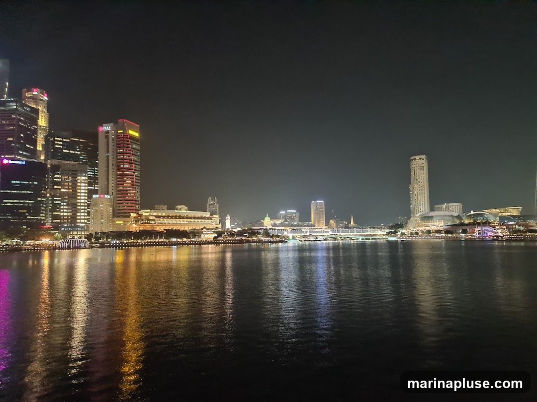 A wide shot of a city skyline at night, with numerous illuminated skyscrapers reflected in the dark water of a bay or river in the foreground. The buildings are a mix of modern and older architecture, with various lights, including colorful ones, visible. The sky is dark and clear.