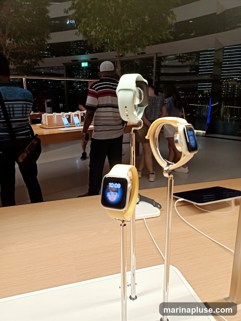 Several Apple Watches are displayed on stands in a store. One watch shows the time 10:09 and a cartoon face. In the background, people are browsing and there are boxes of products on a table. The store appears to have large windows with a city view at night.