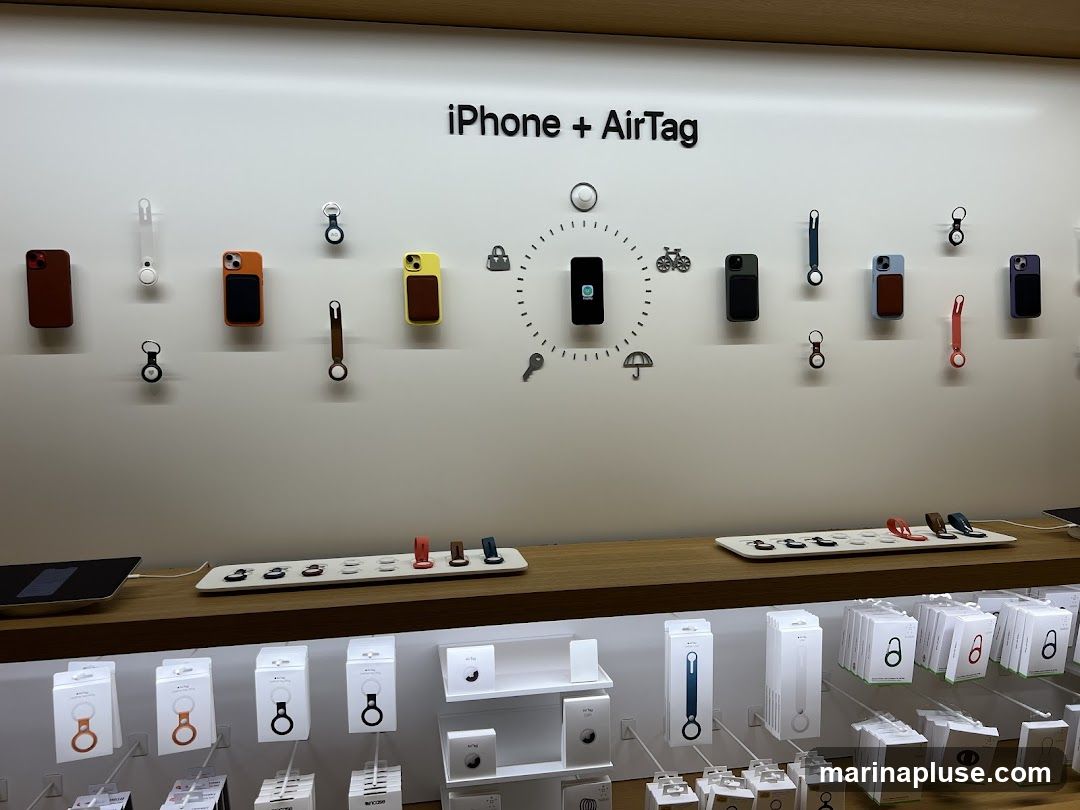 A retail display showcasing various iPhone cases and AirTag accessories. The wall features the text "iPhone + AirTag" and a circular graphic with icons representing a phone, bicycle, lock, key, and umbrella. Various colored iPhone cases and AirTag holders are mounted on the wall. Below, a wooden shelf displays more accessories, and further down, shelves are stocked with boxed products, including AirTags and their accessories.