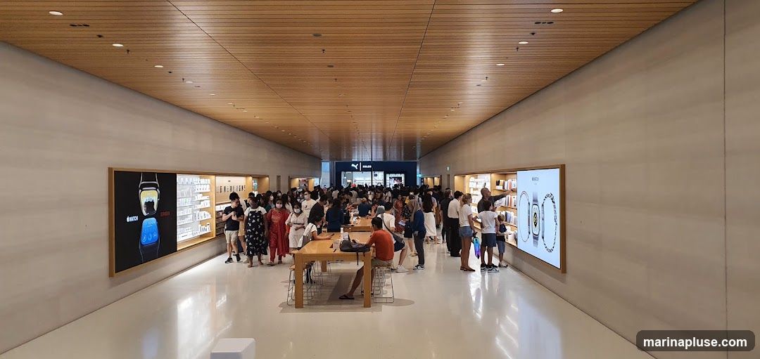 A wide shot of a crowded electronics store with many people browsing. The store has a modern design with wooden ceilings and white walls. Displays on the walls show images of watches and various products. In the background, a PUMA store sign is visible.