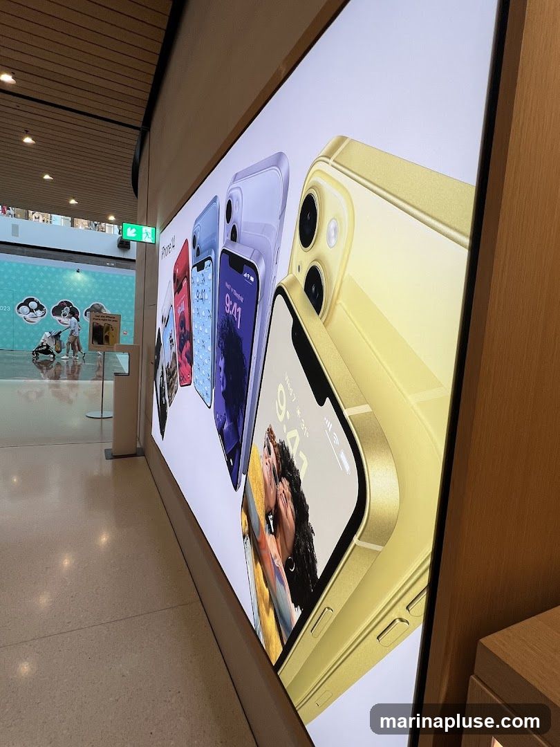 A brightly lit display showcasing several iPhone 14 models in various colors, including yellow, purple, and red. The phones are arranged in a row, with the yellow iPhone 14 prominently featured in the foreground. The display is set against a white background, and the phones show the time and date on their screens. In the background, a green exit sign is visible, along with a mural and people walking in a mall.