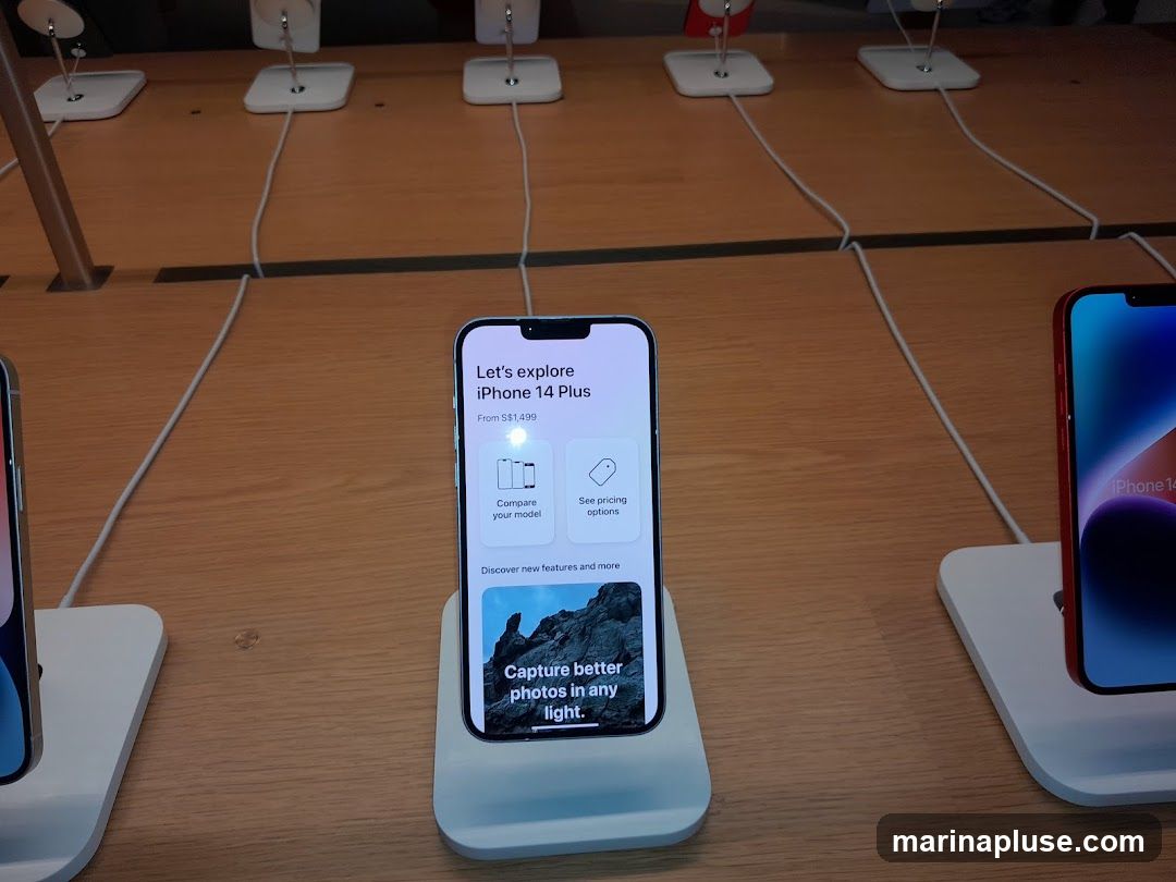 A display of iPhones on a wooden counter. The central phone shows a screen advertising the iPhone 14 Plus, with options to compare models and see pricing. It also highlights the camera capabilities. Another iPhone is visible on the right, displaying "iPhone 14".