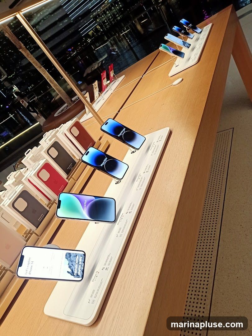 A display table in what appears to be an Apple Store, showcasing various iPhone models and accessories. Several iPhones are arranged on stands, with some displaying promotional information. Cases for iPhones are neatly stacked in their packaging. The background shows a blurred view of a city at night through a large window.