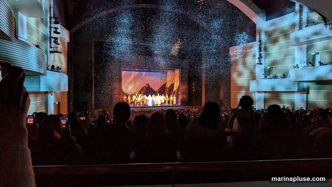 A wide shot of a theatrical performance taking place on a stage. A large group of performers in costume are gathered on stage, with a painted backdrop depicting a mountainous landscape. Confetti or glitter is falling from above, creating a sparkling effect. The audience is seated in the foreground, silhouetted against the stage lights. Balconies with additional audience members are visible on the sides of the theater.