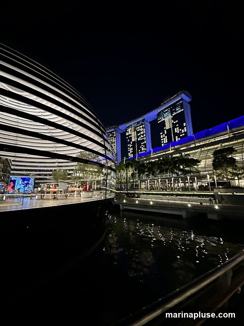 A nighttime view of the iconic Marina Bay Sands hotel in Singapore, with its distinctive three towers connected by a sky park. To the left, a modern, curved building with a striped facade is visible. The scene is illuminated by artificial lights, reflecting on the dark water in the foreground. People are seen in the distance, suggesting a lively atmosphere.