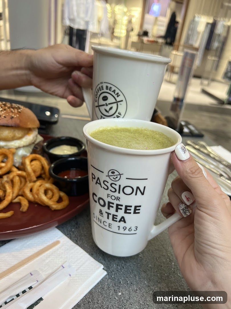 Two hands hold mugs of coffee, one with the logo "The Coffee Bean & Tea Leaf" and the other with "Passion for Coffee & Tea Since 1963." In the foreground, a plate holds a burger and curly fries with dipping sauces. The background appears to be an indoor setting with clothing racks visible.