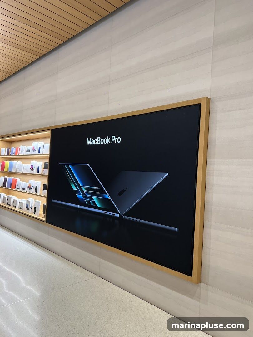 A large screen displays an advertisement for the MacBook Pro, featuring an image of the laptop against a dark background. To the left, shelves are stocked with various Apple products in their packaging. The interior features light-colored walls and a wooden ceiling.