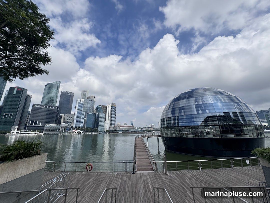 A wide shot of a modern city skyline with a distinctive spherical glass building on the water in the foreground. A wooden walkway leads to the spherical building. The sky is partly cloudy.