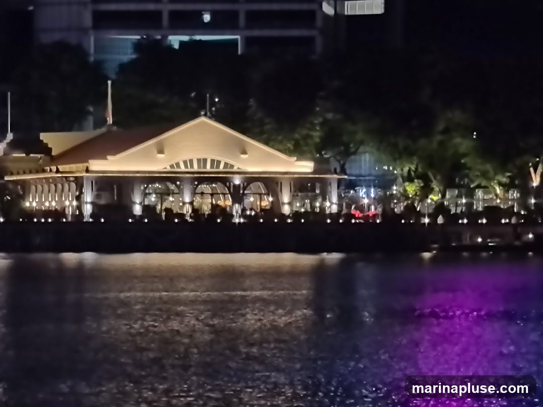 A brightly lit building with a red-tiled roof sits beside a body of water at night. The building appears to be a restaurant or establishment with large windows and outdoor seating. Lights illuminate the facade and the surrounding trees. The water reflects the lights, with a section of purple illumination visible. Silhouettes of people can be seen inside the building.