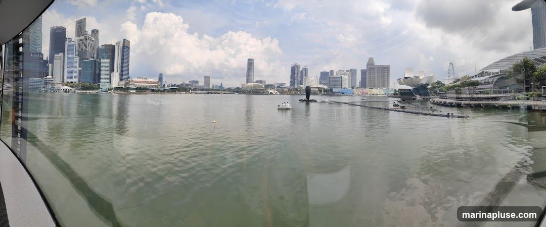 A panoramic view of the Marina Bay skyline in Singapore, with modern skyscrapers and distinctive architectural landmarks such as the Marina Bay Sands and the ArtScience Museum. The water in the foreground reflects the sky and buildings. Several boats are visible on the water, and a Ferris wheel can be seen in the distance. The image appears to be taken from an elevated indoor vantage point, possibly through a window.
