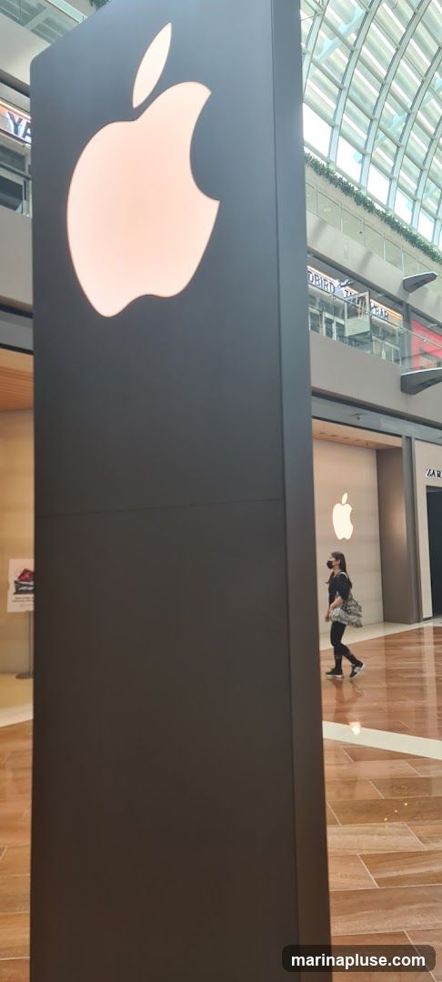 A large, dark pillar with a glowing Apple logo is in the foreground. In the background, the facade of an Apple Store is visible, also featuring a large illuminated Apple logo. A person wearing a mask walks past the store entrance. Other storefronts are visible in the distance.
