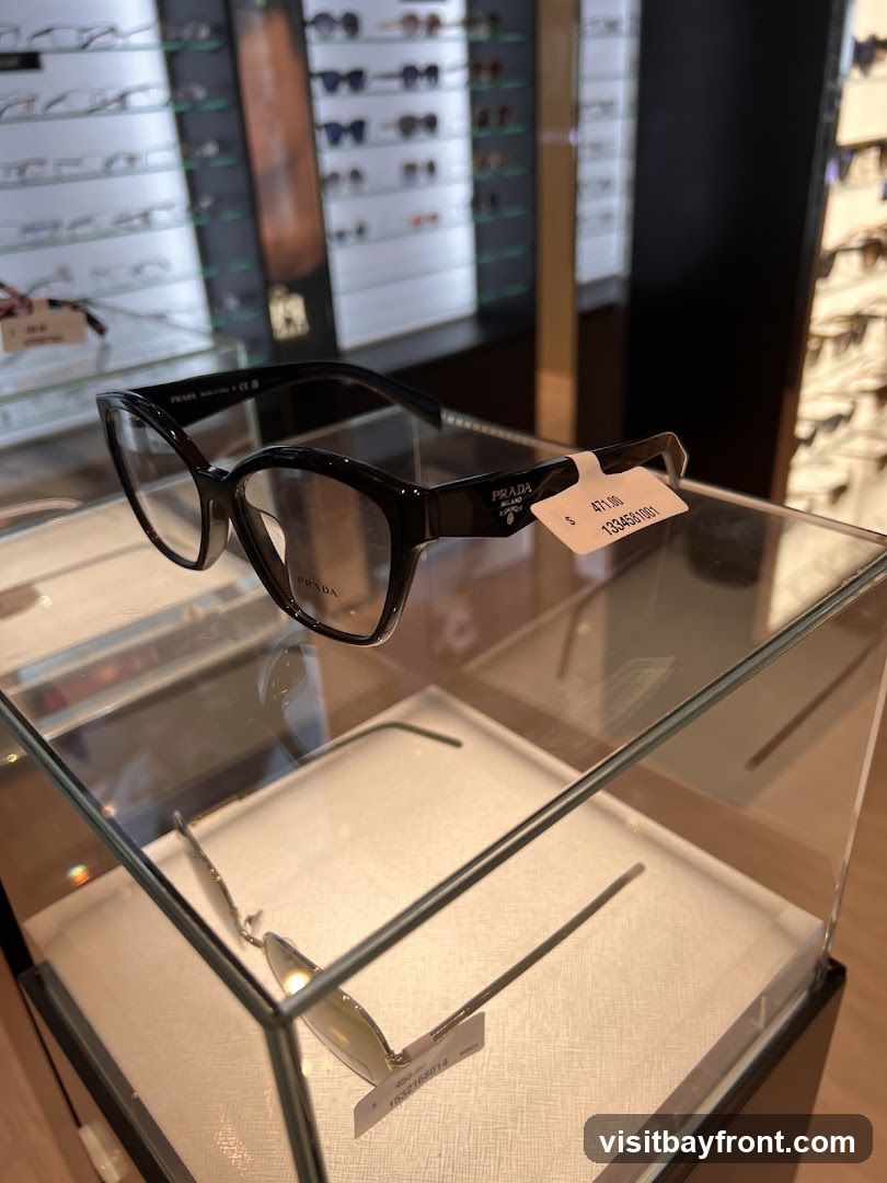 A display case with Prada eyeglasses. The foreground shows a pair of black Prada eyeglasses with a price tag of $471.00. In the background, shelves are filled with various other eyeglasses.