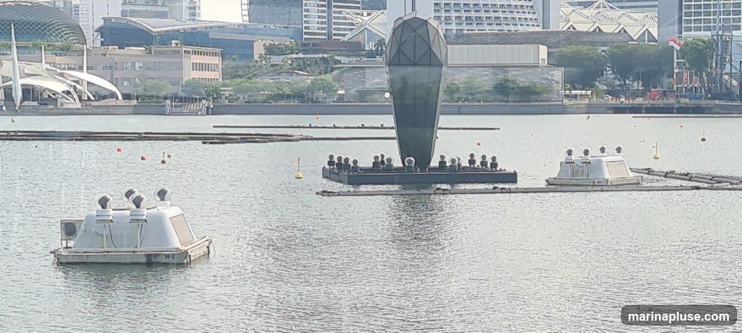 A wide shot of a body of water with several floating structures. In the foreground, a white, boat-like structure with four dome-shaped lights on top is visible. Further out, a larger, dark platform supports a tall, diamond-shaped sculpture, and another smaller white structure. In the background, a cityscape with modern buildings and a distinctive domed structure is seen. The water is calm with ripples.
