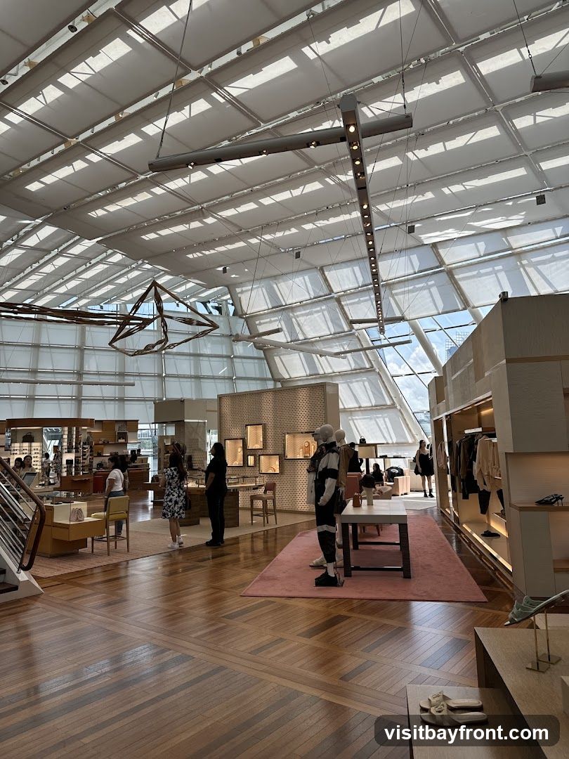 An interior view of a spacious, modern luxury retail store. The store features a high, sloped ceiling with translucent panels that allow natural light to filter in, creating a bright and airy atmosphere. The flooring is polished hardwood, laid in a geometric pattern. Displays of clothing, accessories, and footwear are visible throughout the store, with mannequins showcasing outfits. Customers and staff can be seen browsing the merchandise. The overall aesthetic is clean, minimalist, and upscale.