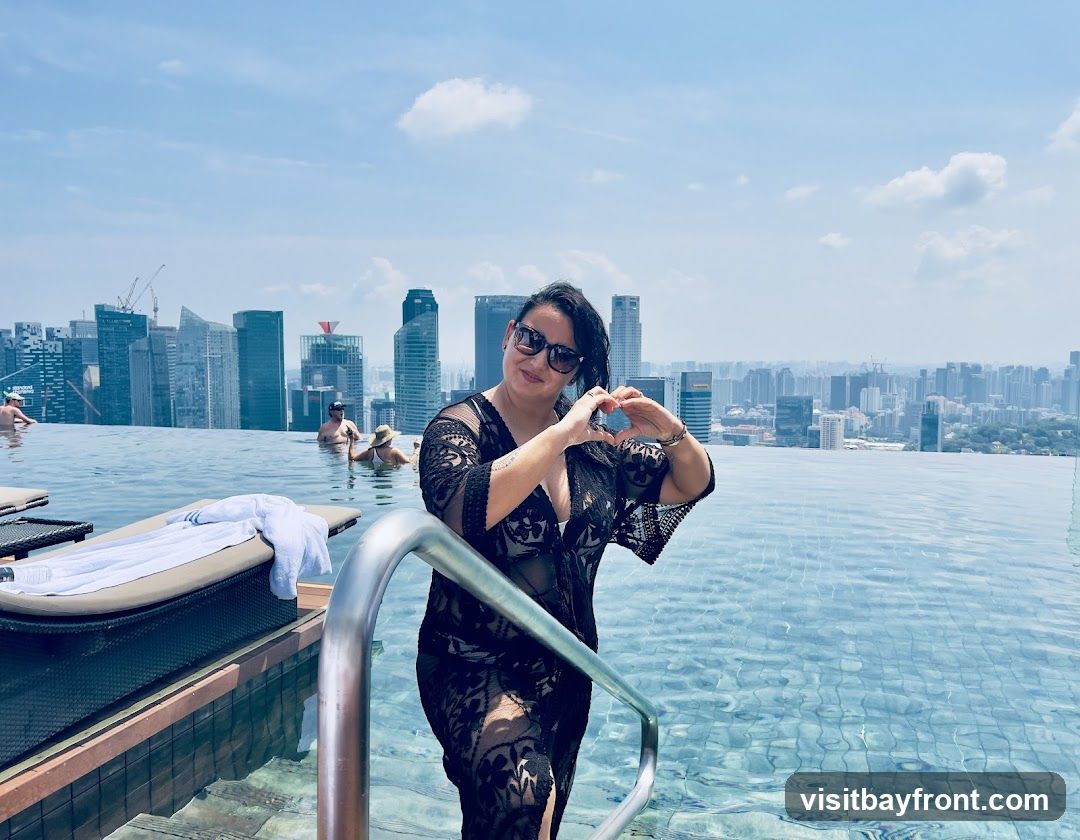 A woman in a black cover-up poses for a photo at an infinity pool, making a heart shape with her hands. She is wearing sunglasses. In the background, a cityscape with numerous skyscrapers is visible under a bright, partly cloudy sky. Other people are in the pool, and lounge chairs are visible at the edge.