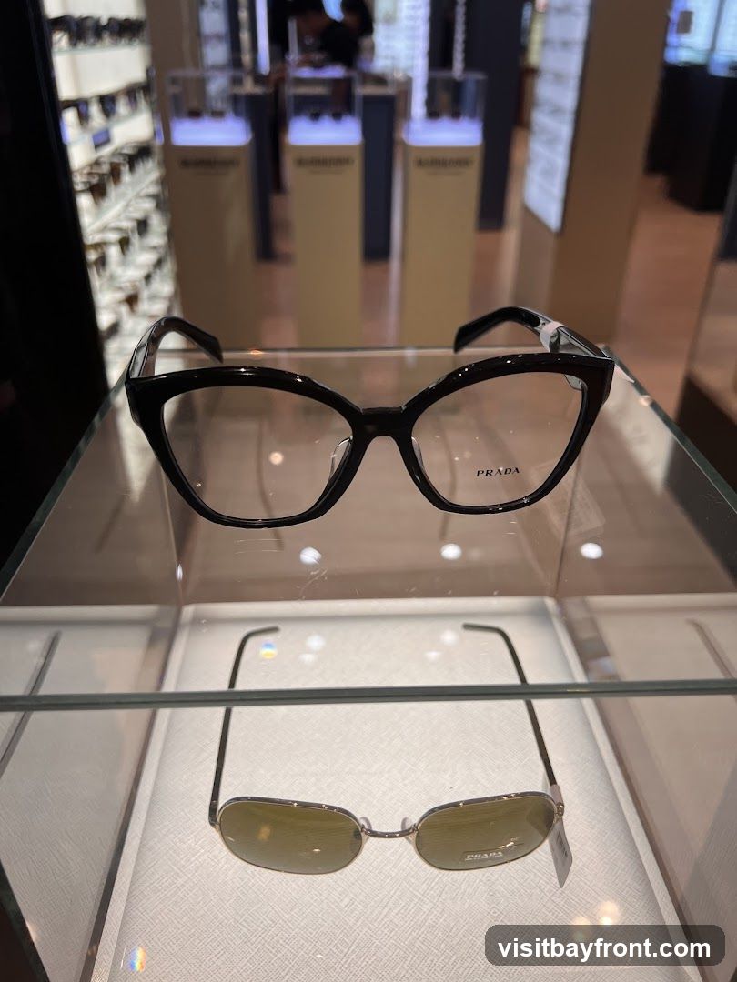 A display case shows a pair of black Prada eyeglasses on the top shelf and a pair of sunglasses on the bottom shelf. The eyeglasses have a cat-eye shape and the Prada logo is visible on the right lens. The sunglasses have a rectangular shape and a tag with the Prada logo is attached to the right temple. In the background, other eyewear is displayed on shelves and illuminated stands.