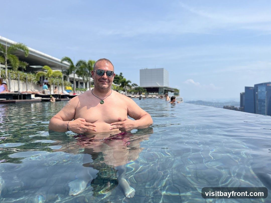 A man poses for a photo in an infinity pool overlooking a city skyline. He is wearing sunglasses and a necklace. Other people are visible in the pool and in the background, along with palm trees and buildings. The sky is blue with some clouds.