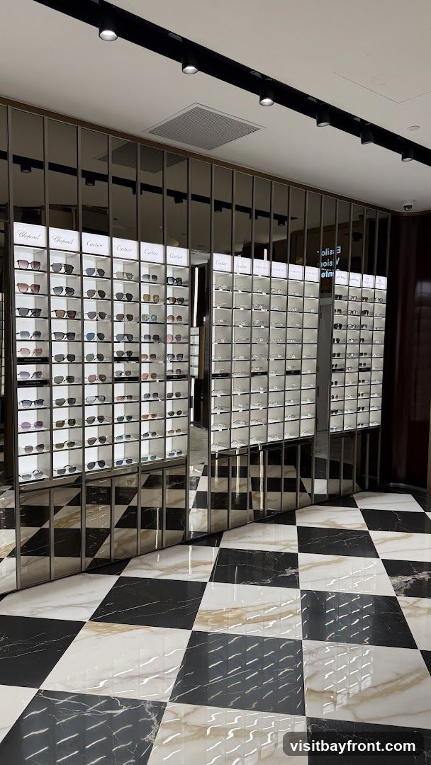 A well-lit interior display of sunglasses and eyeglasses, organized by brand in illuminated shelves. Brands visible include Chopard and Cartier. The floor features a striking black and white checkered marble pattern.