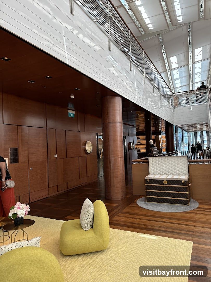 An interior view of a high-end retail store, likely a luxury brand, featuring a large, ornate trunk displayed prominently on a circular rug. The store has a modern, spacious design with polished wooden floors and walls. A mezzanine level is visible with railings and people browsing. Seating areas with modern, colorful furniture are arranged on a textured yellow carpet. An 'EXIT' sign is visible on the wood-paneled wall.