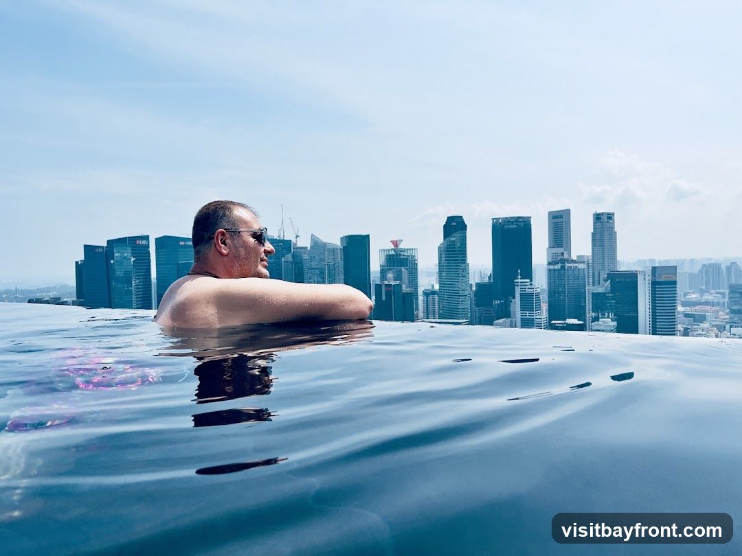 A man relaxes in an infinity pool, with a panoramic view of a city skyline in the background. The water in the foreground is calm, with gentle ripples. The man is wearing sunglasses and looking to the right. The skyscrapers in the distance are tall and modern, suggesting a bustling urban environment. The sky is clear and blue with a few scattered clouds.