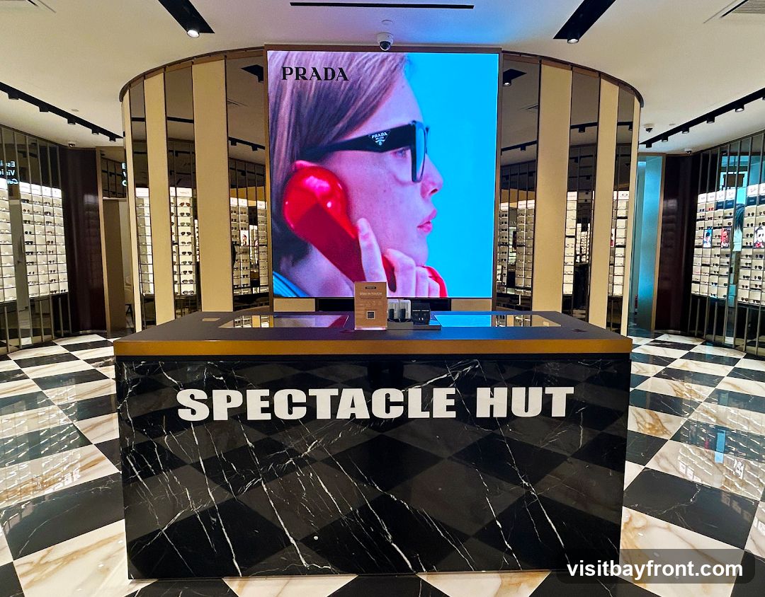 The interior of a modern optical shop with a checkered floor. A large digital screen displays a Prada advertisement. In the foreground, a black marble counter with the words "SPECTACLE HUT" in white lettering is visible. The walls are lined with display shelves filled with eyeglasses.