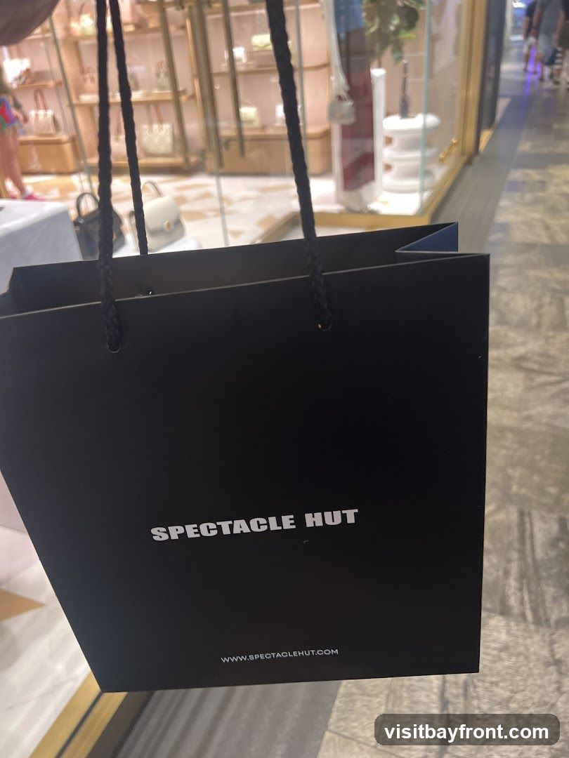 A black shopping bag with the words "SPECTACLE HUT" and "WWW.SPECTACLEHUT.COM" printed on it. The bag is held by its handles, and in the background, a store window displays various handbags.