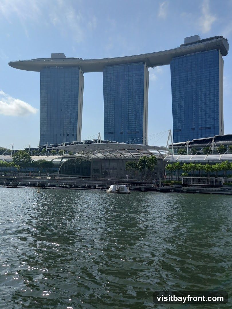 A wide shot of the Marina Bay Sands complex in Singapore, with its iconic three towers connected by a sky bridge. The towers are tall and modern, with blue glass facades. In front of the towers is a lower, curved building with a glass facade and a white roof structure. The foreground shows a body of water with ripples reflecting the sunlight. A small white boat is visible on the water. The sky is blue with some scattered clouds.