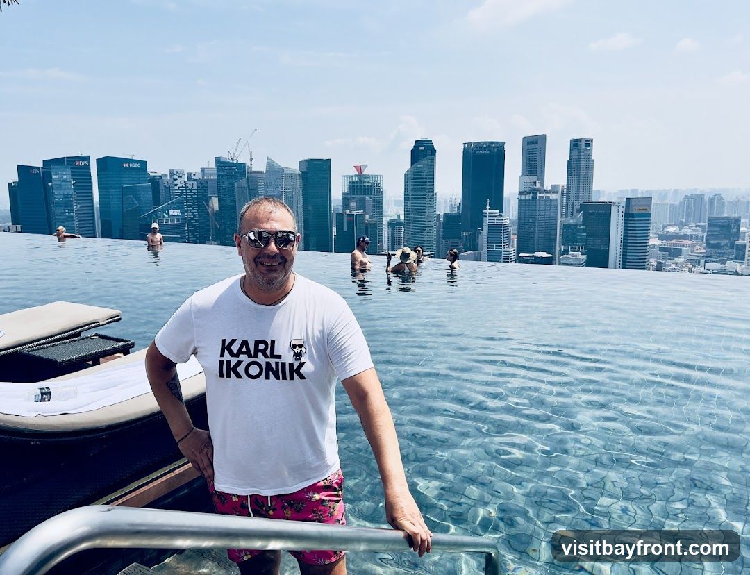 A man wearing a white t-shirt with the text "KARL IKONIK" and pink patterned shorts stands in an infinity pool. He is smiling and wearing sunglasses. In the background, a dense city skyline with numerous skyscrapers is visible under a bright, hazy sky. Several other people are also in the pool.