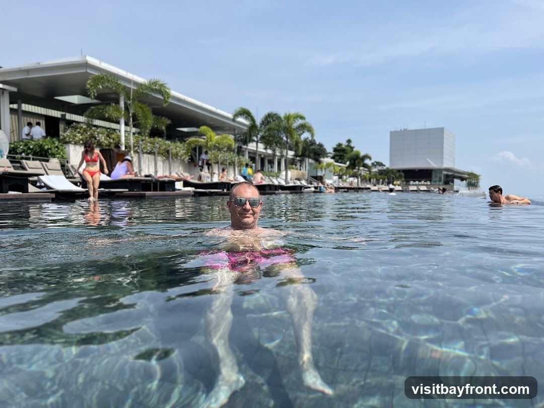 A man is swimming in an infinity pool with lounge chairs and palm trees in the background. The sky is blue with some clouds.
