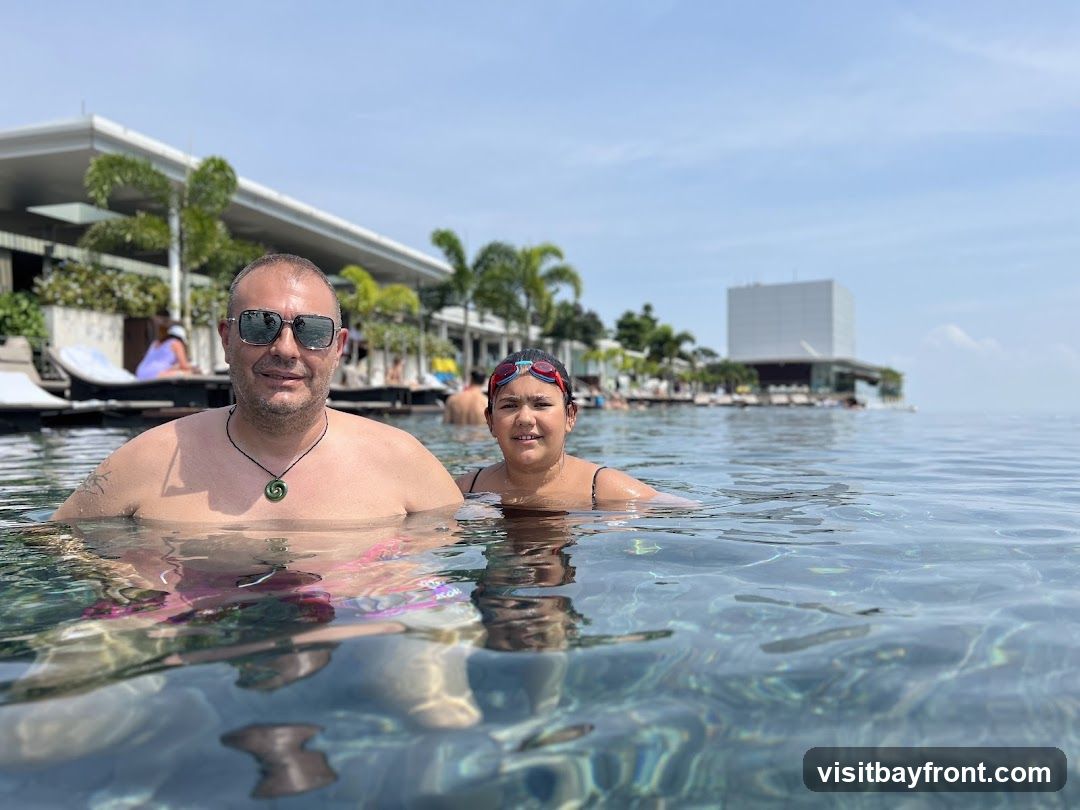 A man and a young girl are in an infinity pool, with a modern building and palm trees in the background. The water is clear and reflects the sky.