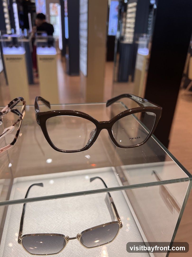 A close-up view of a pair of brown Prada eyeglasses displayed on a glass shelf in an optical store. Below them, a pair of gold-rimmed sunglasses with gradient lenses is also visible. In the blurred background, other eyewear displays and a person can be seen.