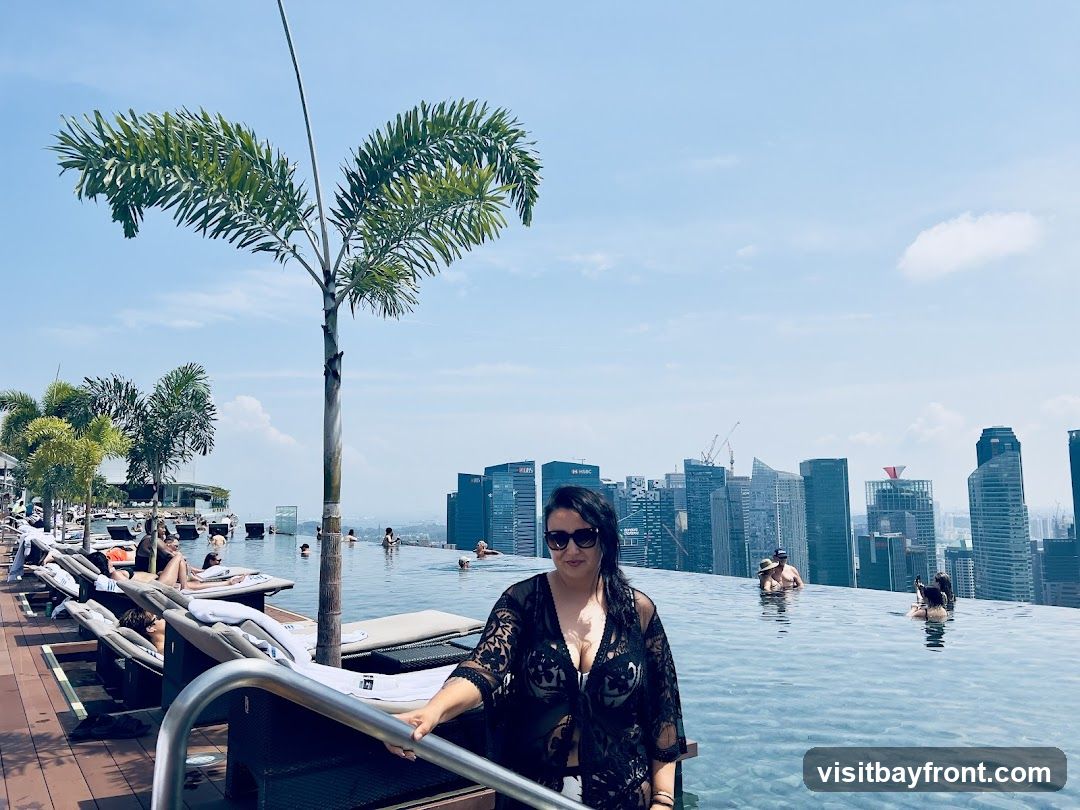 A woman in a black lace cover-up stands by the edge of an infinity pool, looking towards the camera. She is wearing sunglasses. In the background, several people are swimming and lounging by the pool. The pool overlooks a dense cityscape with numerous skyscrapers under a clear blue sky. Palm trees are visible on the left side of the image.
