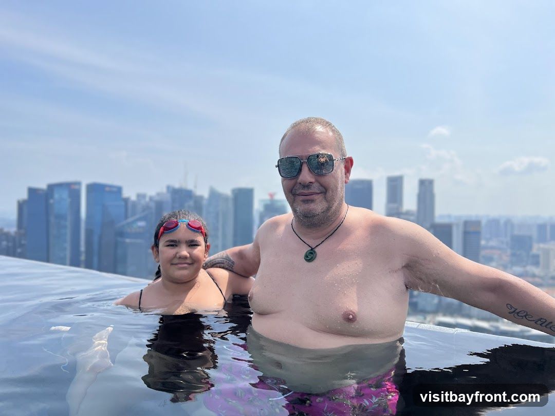 A man and a young girl are in an infinity pool, with a cityscape in the background. The man is wearing sunglasses and a necklace. The girl is wearing swimming goggles. The water is calm and reflects the sky and buildings.