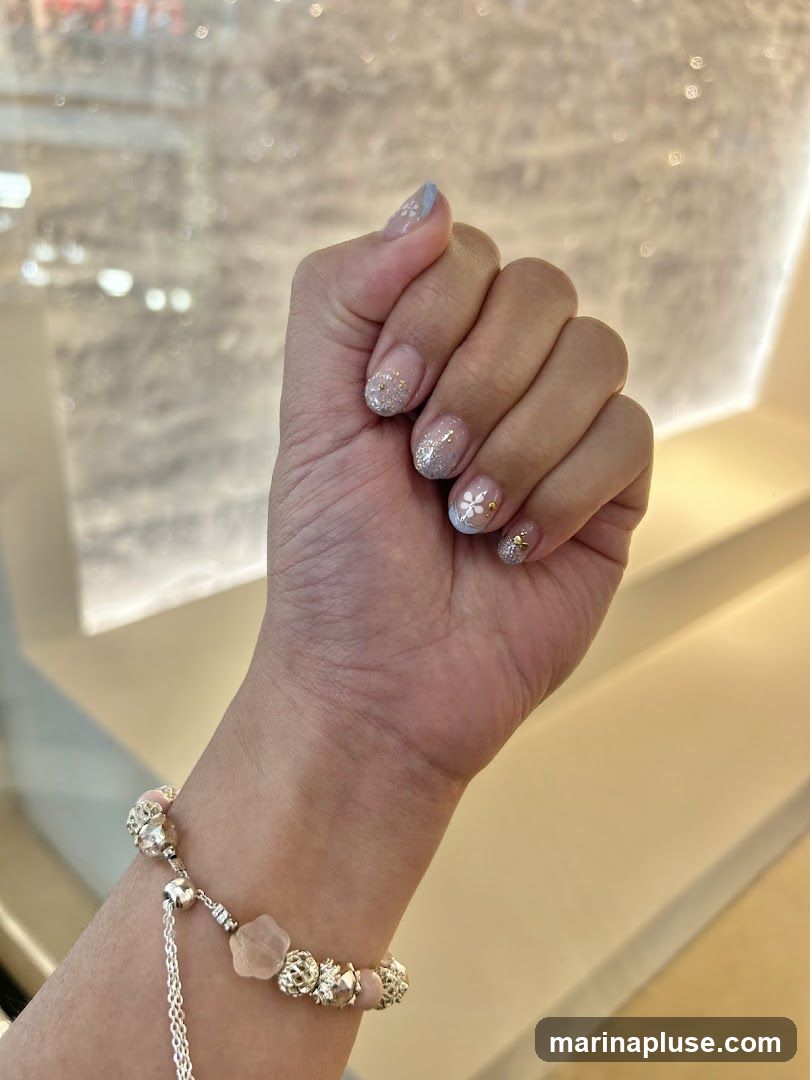 A close-up shot of a person's hand displaying intricate nail art with glitter and floral designs. The nails are painted with a light base color, adorned with small white flowers and sparkling glitter. A delicate bracelet with pink beads and silver charms is worn on the wrist. The background is softly blurred, suggesting an indoor setting with warm lighting.