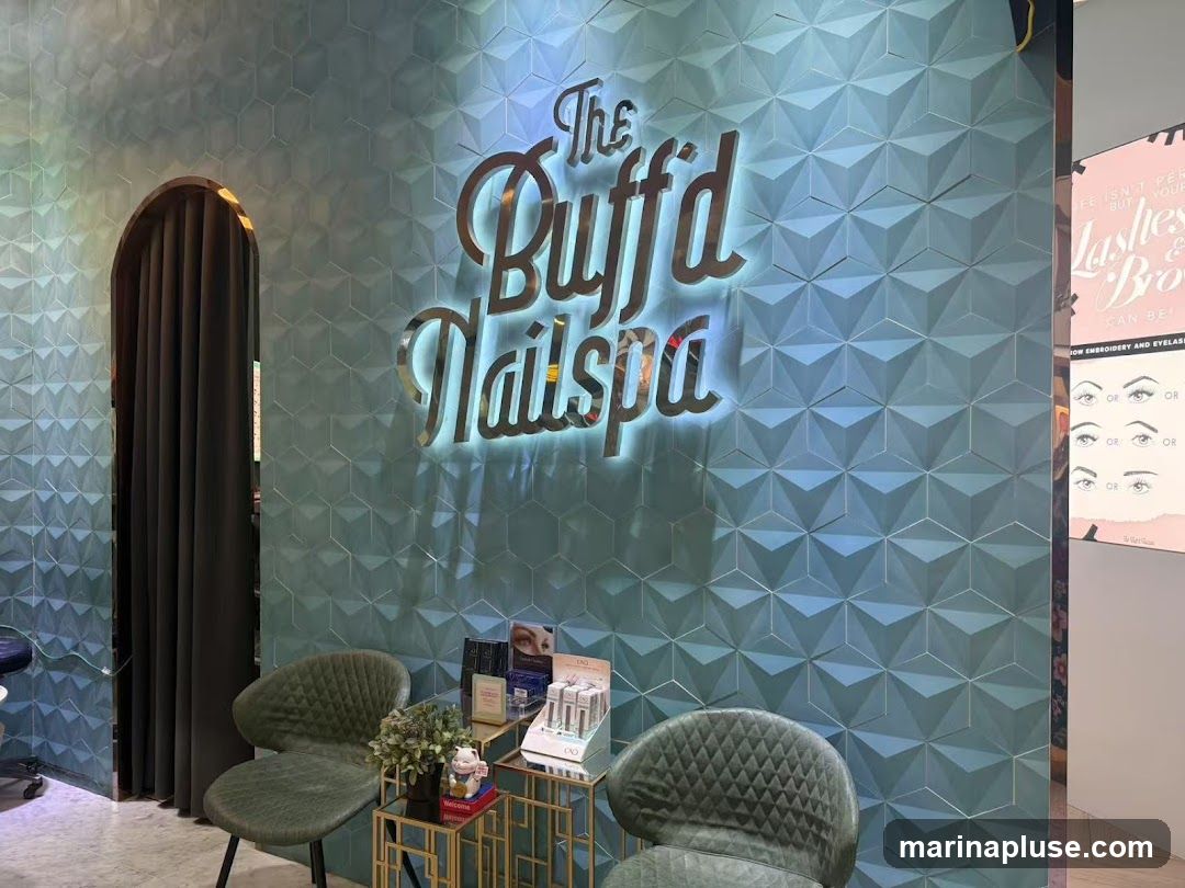 The interior of a nail spa with a textured blue wall featuring the illuminated sign "The Buff'd Nailspa". Two green chairs are placed in front of the wall, with a small table displaying various beauty products and a decorative item. To the right, a poster advertises eyelash and eyebrow services. An arched doorway with dark curtains is visible on the left.
