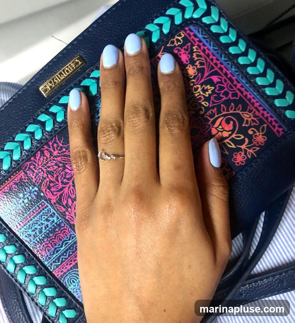 A close-up shot shows a person's left hand with light blue nail polish. The hand is resting on a dark blue clutch with a colorful, embroidered pattern featuring floral and paisley motifs in shades of pink, orange, blue, and purple. The clutch also has decorative teal stitching along the edges. A thin, silver ring with a crescent moon design is worn on the ring finger. The background appears to be a striped fabric, possibly a shirt or bag lining.