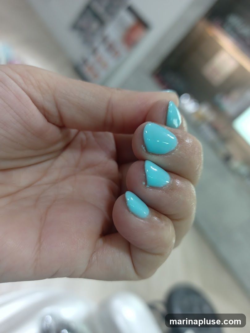 A close-up shot focuses on a person's hand, showcasing freshly painted turquoise nails. The hand is positioned in the foreground, with the nails appearing smooth and glossy. In the blurred background, there are hints of what appears to be a salon or retail environment, with shelves or displays visible. The lighting suggests an indoor setting.