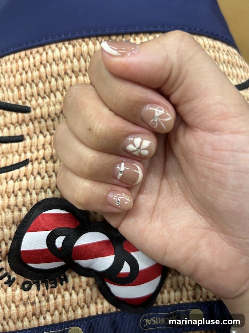 A close-up shot of a person's hand with manicured nails. The nails have a nude base with white French tips. Some nails are decorated with white floral designs and small rhinestones, while others feature a white bow design with a rhinestone in the center. The hand is resting on a straw-like textured surface with a blue trim and a Hello Kitty bow emblem.
