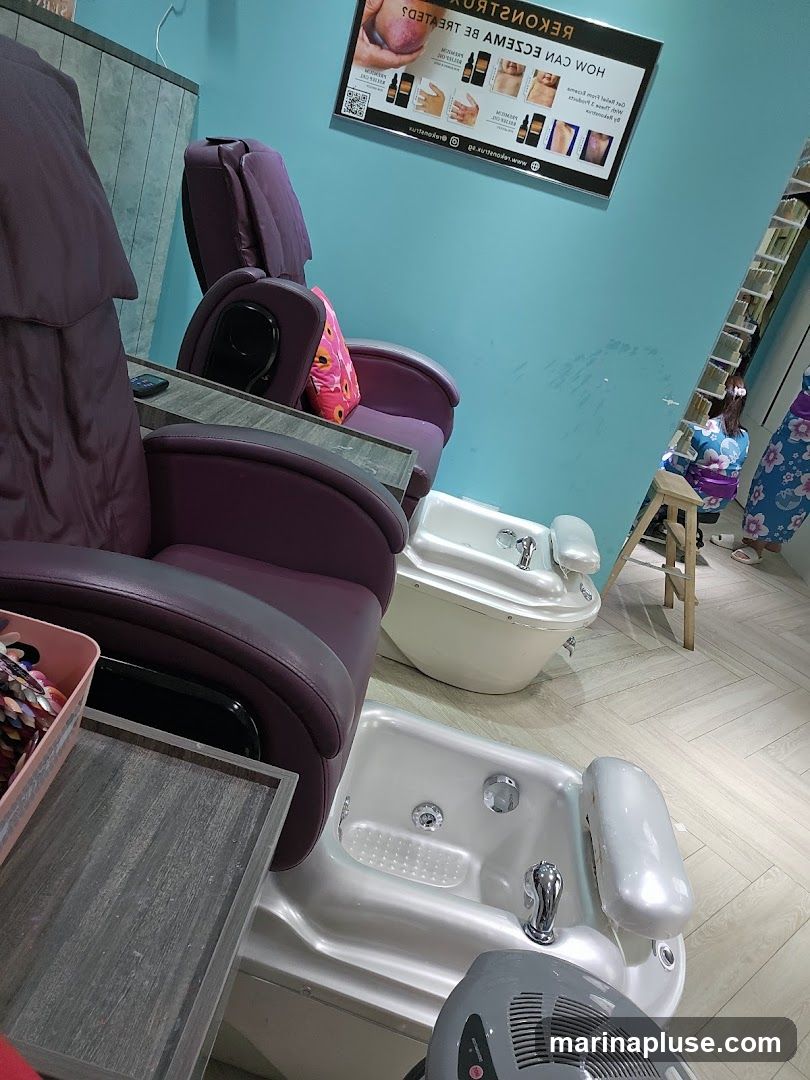 A salon interior featuring two plush purple pedicure chairs with built-in side tables. In front of each chair is a white foot bath with chrome fixtures. A poster on the teal wall displays information about treating eczema. In the background, a person in a kimono-like garment is visible near a shelf unit. A pink basket filled with nail art supplies sits on a side table.