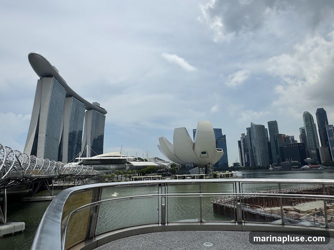 A wide shot of the Singapore skyline featuring the iconic Marina Bay Sands hotel, the ArtScience Museum, and other skyscrapers. The view is from a waterfront promenade with a railing in the foreground. The sky is cloudy.