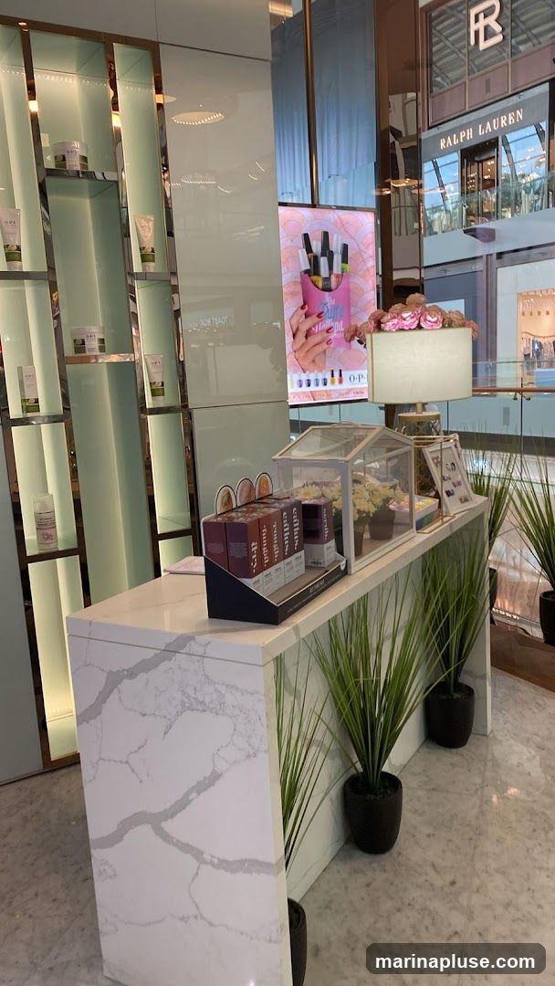 A display counter with various beauty products, including OPI nail polish and skincare items. There is a digital screen showing a hand holding nail polish bottles. The counter is decorated with artificial plants and a floral arrangement. In the background, a Ralph Lauren store is visible.