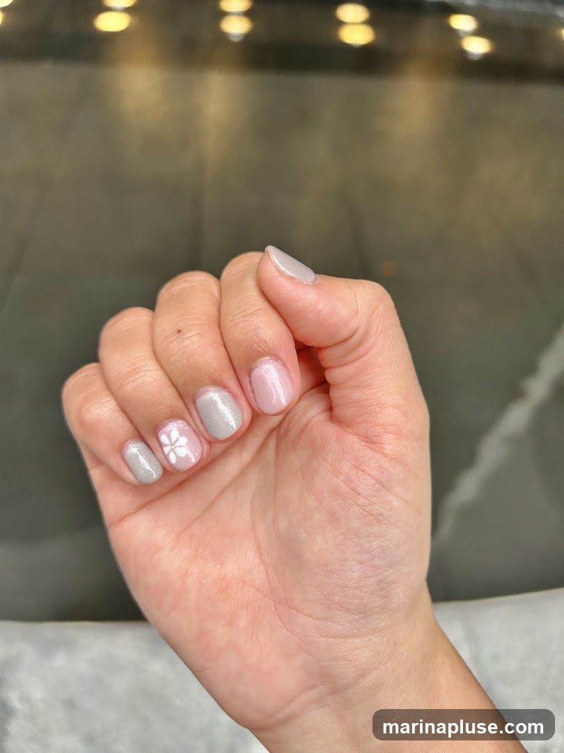 A close-up of a hand with light pink and grey nail polish. Some nails have a subtle glitter finish, while one nail features a delicate white flower design. The background is blurred, showing warm, out-of-focus lights.