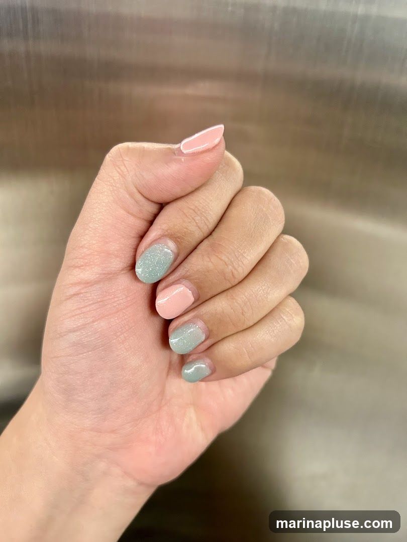 A close-up shot of a hand with manicured nails. The nails are painted in alternating colors of pale pink and a light, shimmery mint green. The thumb and index finger have pale pink polish, while the middle and ring fingers have the mint green polish. The pinky finger is not fully visible. The background is a blurred, metallic surface.