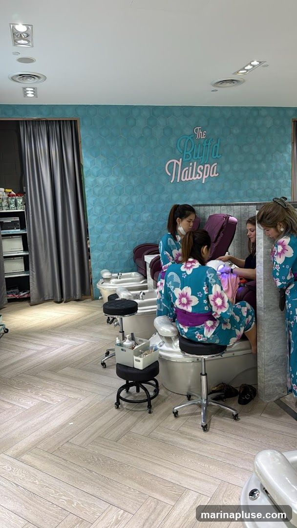 A nail spa interior with several pedicure stations. Three staff members, wearing matching floral kimonos, are attending to clients. One client is seated at a pedicure station, receiving nail treatment. Another client is seated nearby. The walls are decorated with a teal hexagonal tile pattern, and a sign reads "The Buff'd Nailspa". Shelving units with products are visible on the left side of the image.