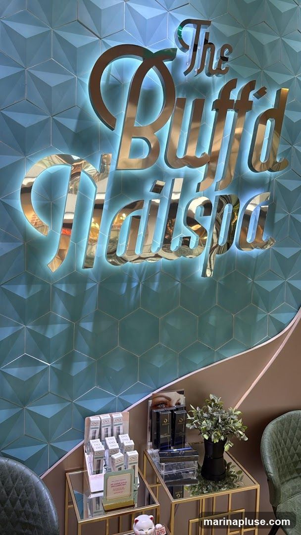 A close-up shot of the illuminated signage for "The Buffd Nailspa" mounted on a textured teal wall. Below the sign, on mirrored tables, are displays of nail care products and a small potted plant.