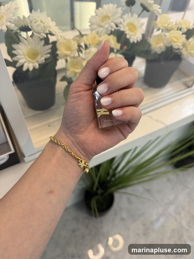 A close-up shot of a woman's hand holding a bottle of nail polish. Her nails are painted with a gradient of white and pink, with a subtle shimmer. She is also wearing a gold bracelet with a charm. In the background, there are artificial white and yellow flowers in dark pots, and a glimpse of a green plant. The overall impression is one of beauty and self-care.