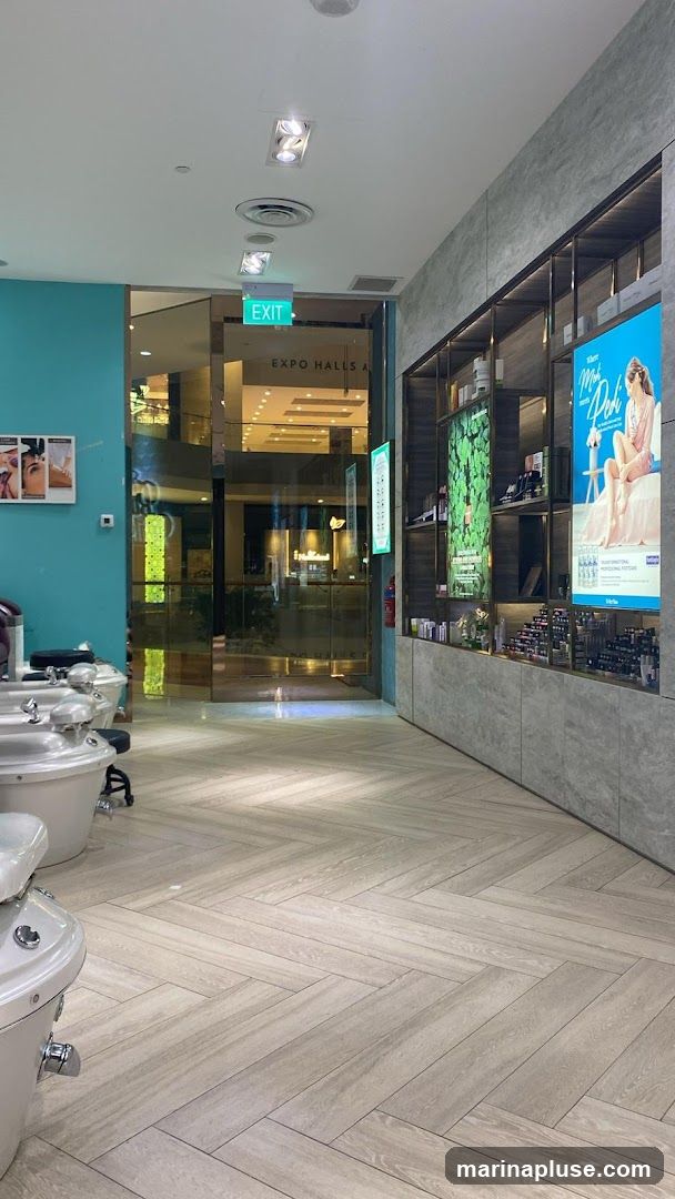 The image shows the interior of a nail salon with pedicure chairs visible in the foreground. The salon features a modern design with light wood-patterned flooring and teal walls. Shelves display various beauty products and promotional posters. In the background, a glass entrance leads to a shopping area with an "EXIT" sign and "EXPO HALLS A" visible.
