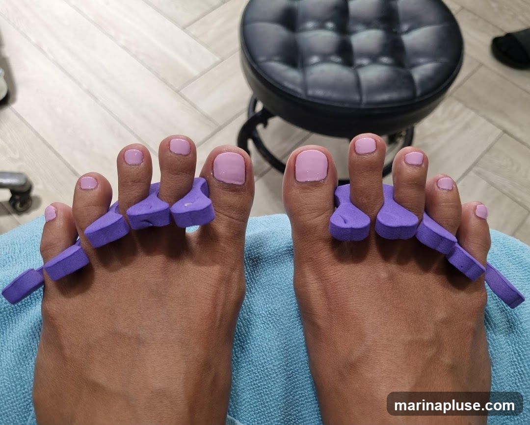 A close-up view of a person's feet with purple toe separators in place, ready for a pedicure. The toenails are painted a light pink color. In the background, a black padded stool is visible, suggesting a salon or spa setting. The floor has a light-colored wood-grain pattern.